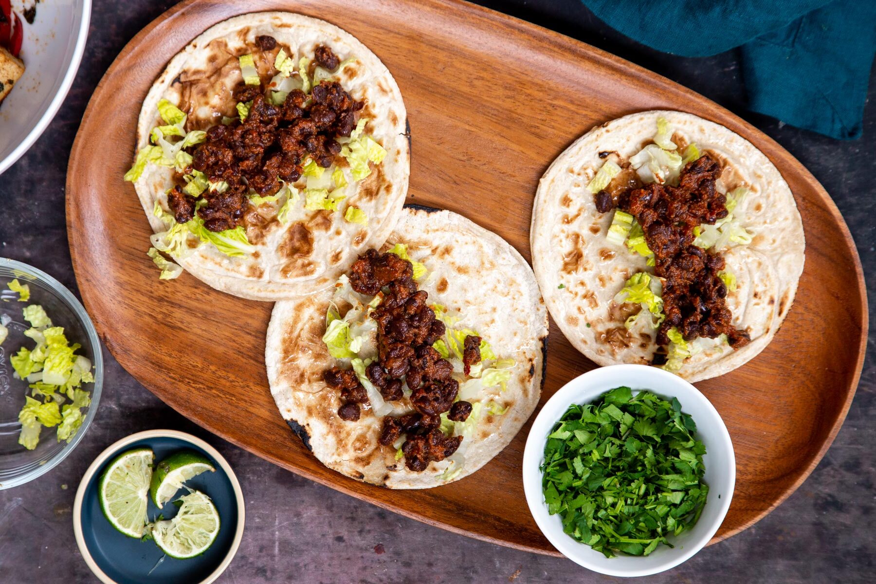 three small wheat tortillas on a wooden platter being topped with lettuce and black beans 