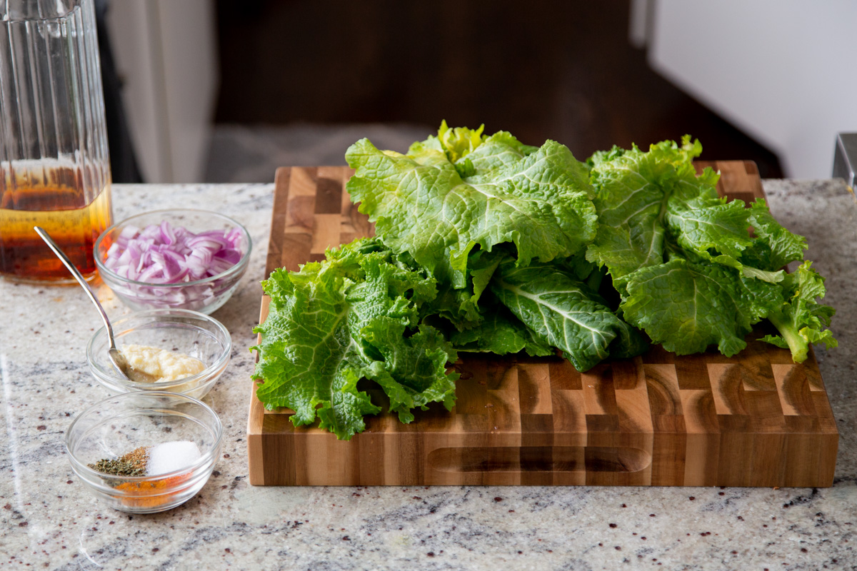 mustard greens on a wooden chopping board with spices and chopped onions in small dishes to one side