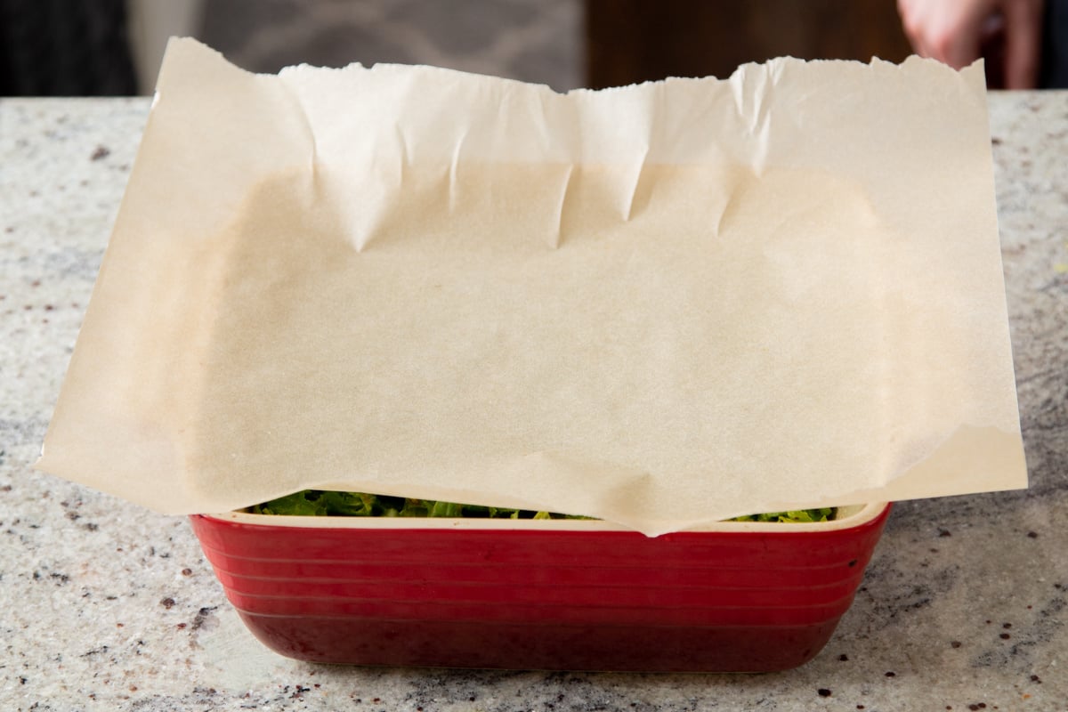 a casserole dish with mustard greens topped with a piece of parchment paper