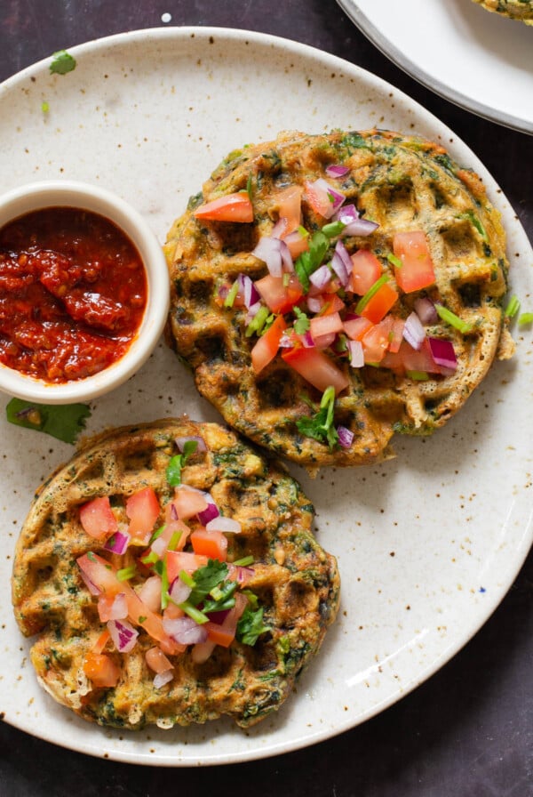 two spinach waffles on a plate topped with chopped tomato and cilantro with red dipping sauce next to them